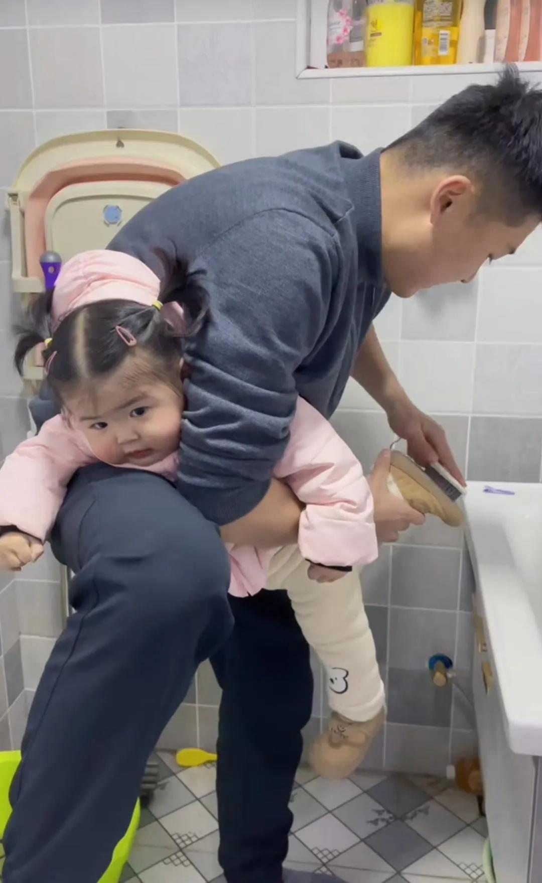 这是网上流传的一个可爱的小女孩被爸爸夹抱着刷鞋的情景。小女孩脸蛋圆圆的，胖乎乎的