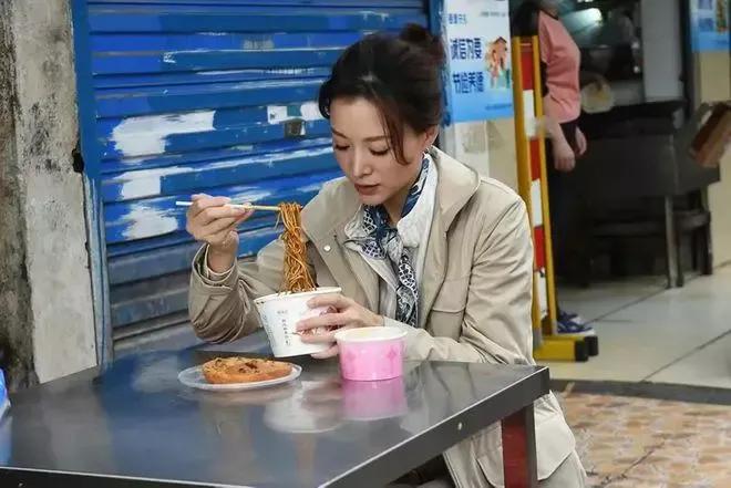 董卿在武汉路边小吃摊上吃热干面，气质优雅，非常接地气。
穿着一身米色风衣，系着蓝